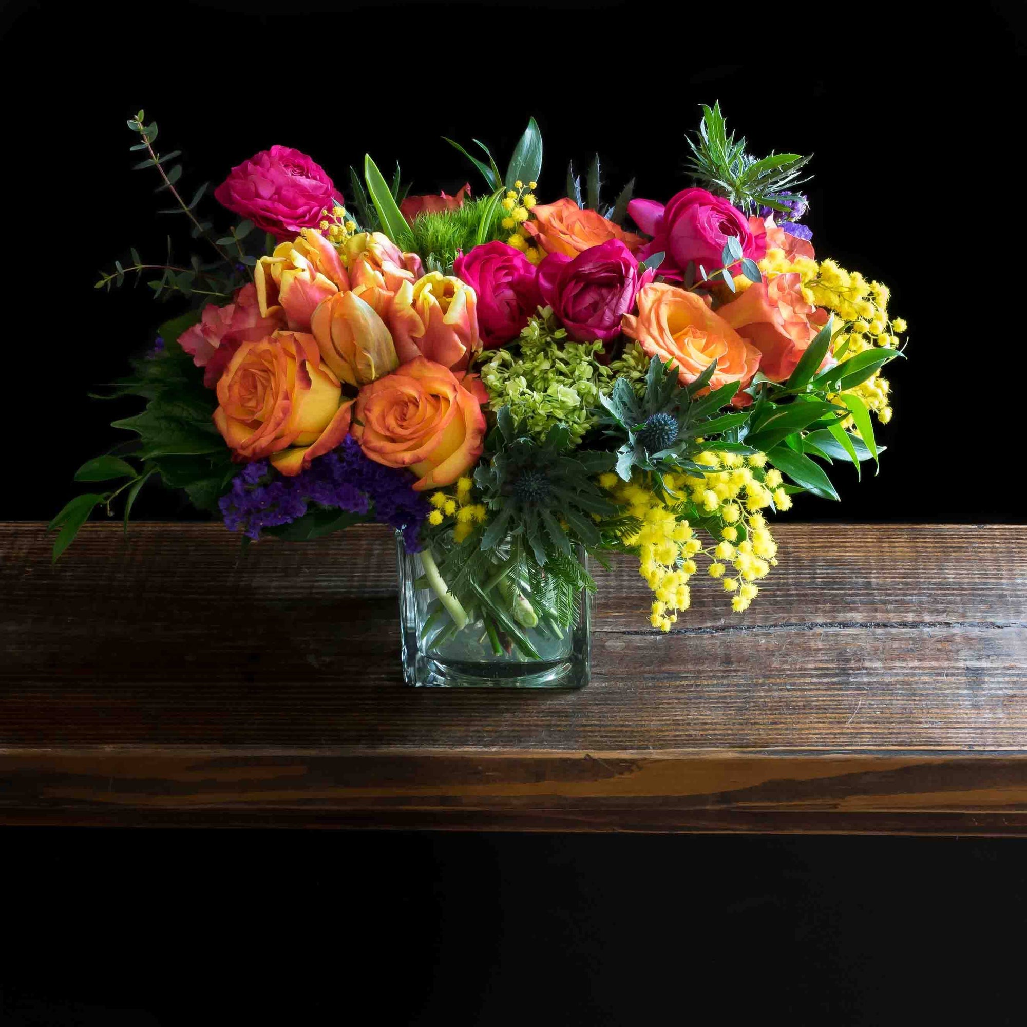 Beautiful boutique colorful floral arrangement with pink ranunculus, yellow and orange tulips, orange roses, purple flowers, and mini green hydrangeas.