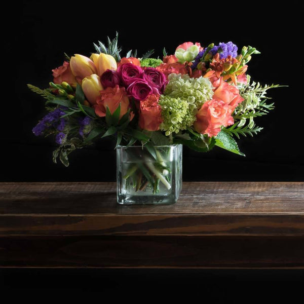 Beautiful boutique colorful floral arrangement with pink ranunculus, yellow and orange tulips, orange roses, purple flowers, and mini green hydrangeas.