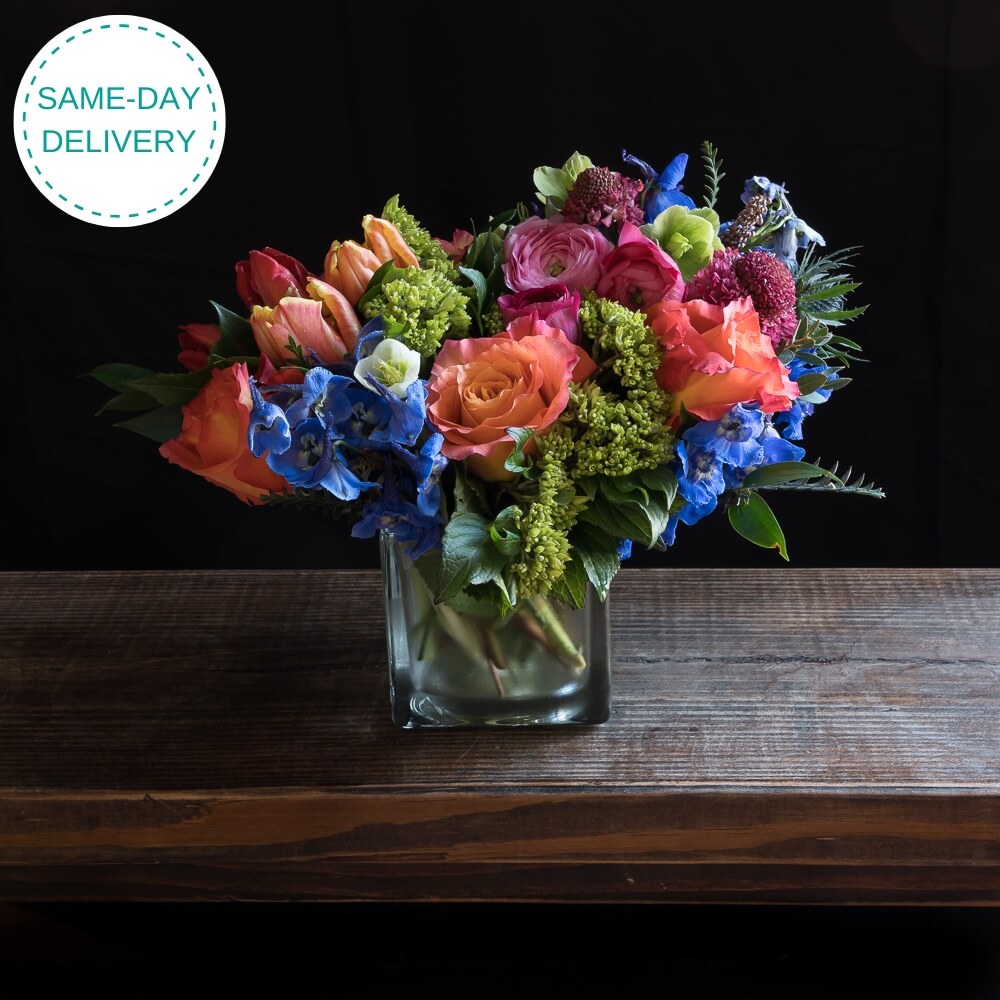 Beautiful boutique colorful floral arrangement with pink ranunculus, yellow and orange tulips, orange roses, purple flowers, and mini green hydrangeas.
