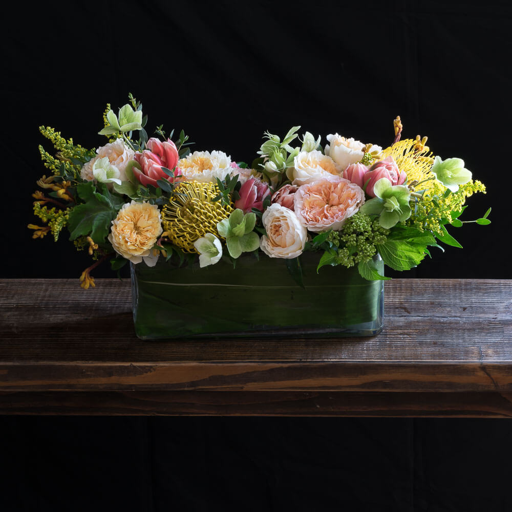 Colorful centerpiece floral arrangement with light pink, yellow roses, cones, and light yellow protea