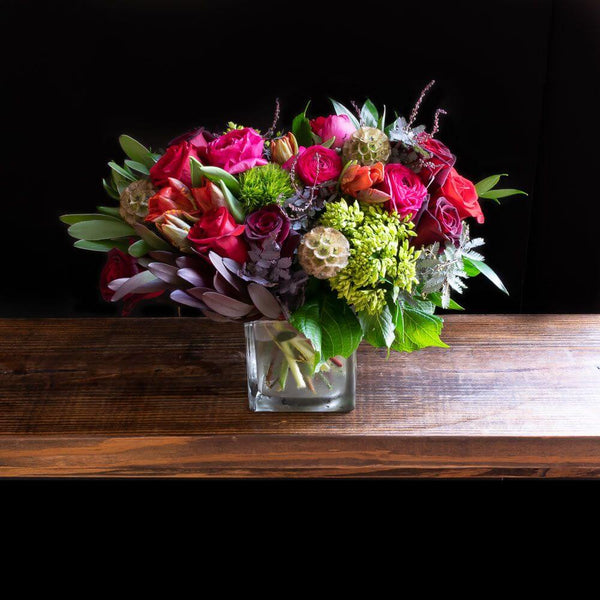 Boutique flower arrangement with red roses, dark pink ranunculus, red parrot tulips, and dark red roses.