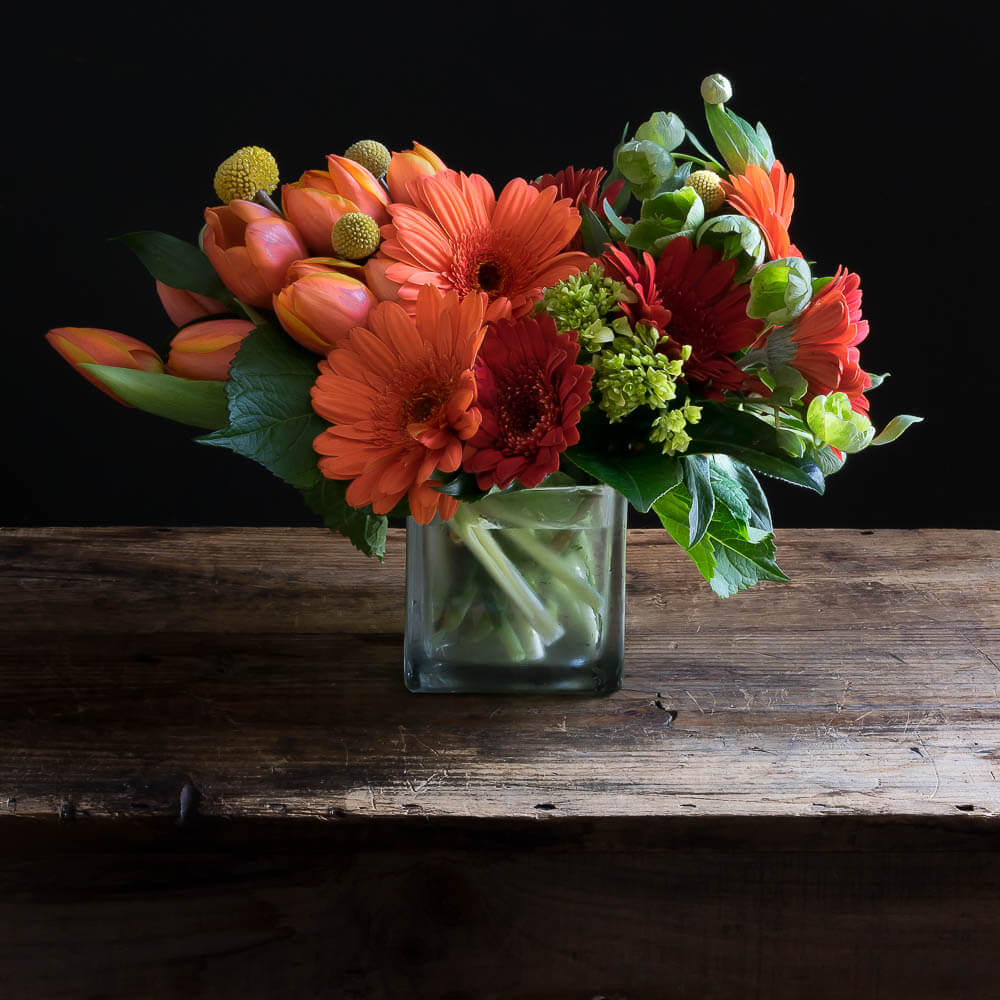 Floral arrangement with orange tulips, orange gerberas, red gerberas, and mini green hydrangeas.