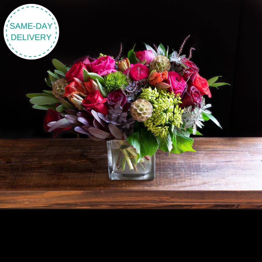 Boutique flower arrangement with red roses, dark pink ranunculus, red parrot tulips, and dark red roses.